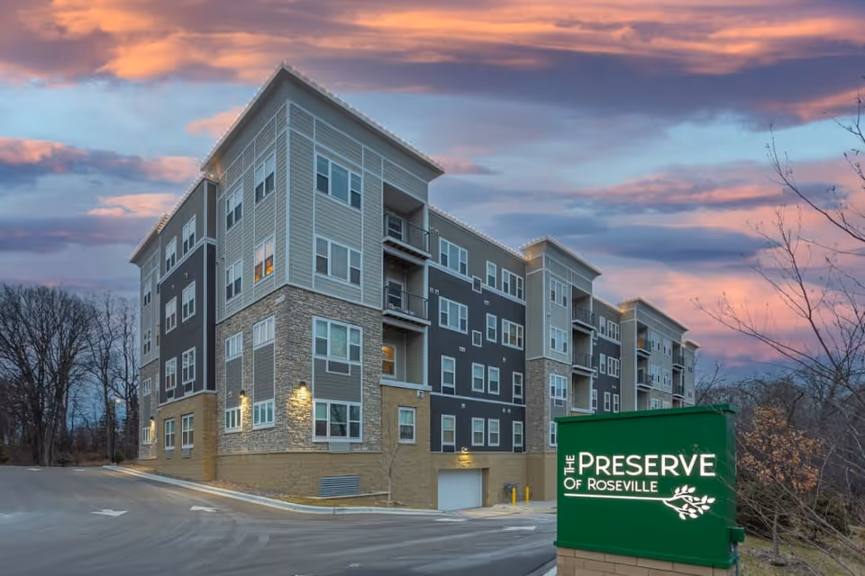 Exterior view of a multi-story senior living building at dusk with a green 'The Preserve of Roseville' sign in front.