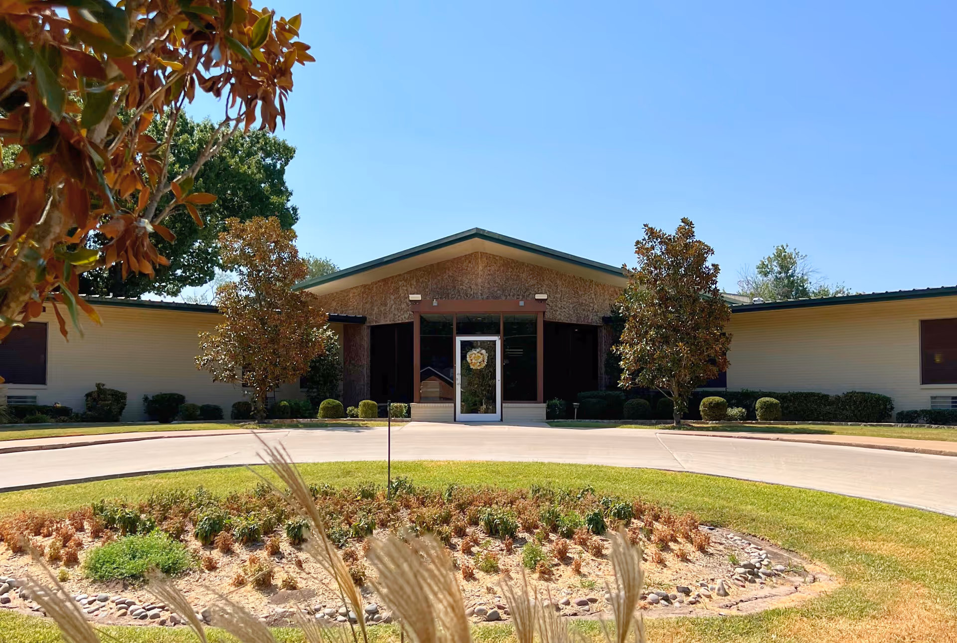 Front entrance of a single-story nursing and rehab building with a circular driveway, landscaped roundabout, and trees.