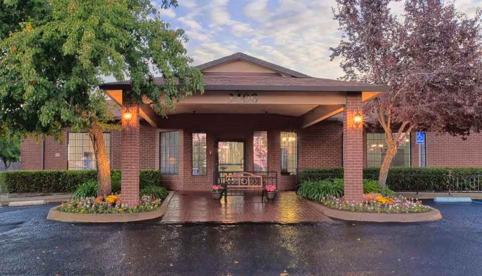 Front entrance of Auburn Oaks Care Center featuring a covered porch supported by brick columns, a bench with flower pots, surrounded by landscaped flower beds and trees, with a wet driveway in front and a partly cloudy sky above.