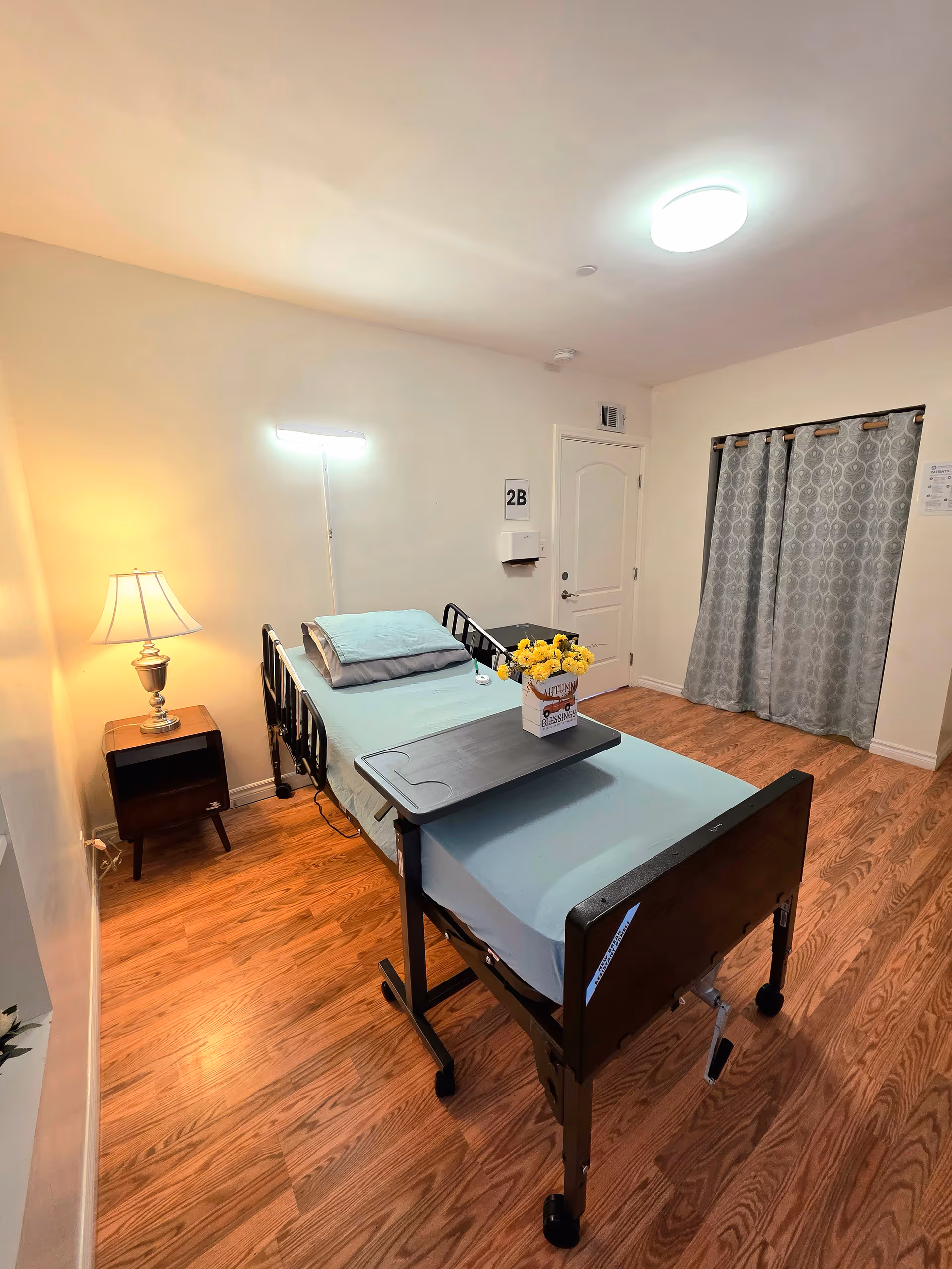 A hospital-style bed in a small bedroom with lamp, flowers, and curtains.