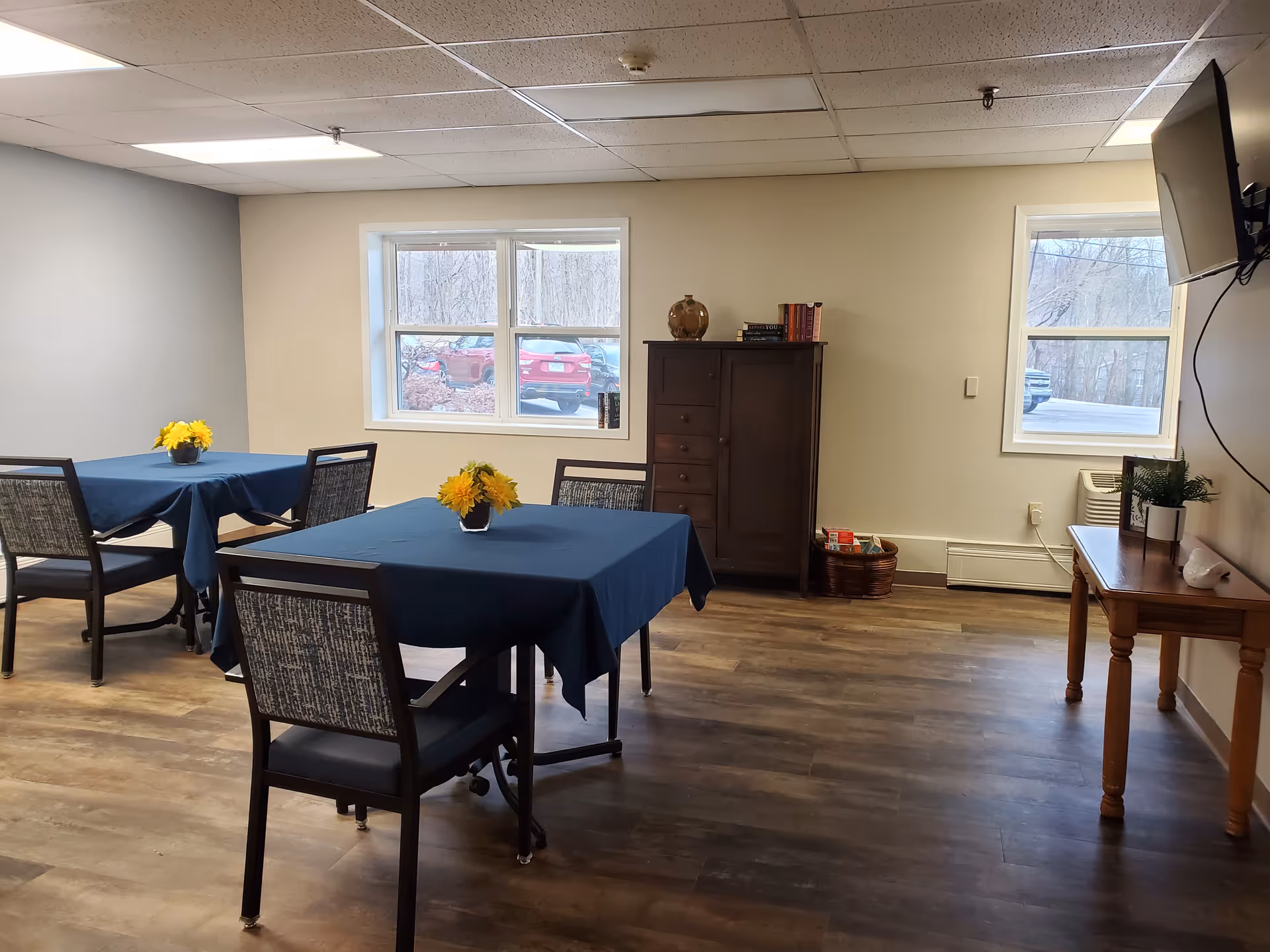 Senior living dining room with blue tablecloths, yellow flowers, and a dark wood cabinet.