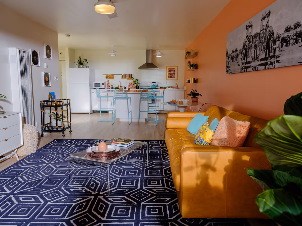 Open-plan living room and kitchen with a mustard leather sofa against an orange wall, geometric rug, clear coffee table, and a kitchen island with turquoise bar stools.