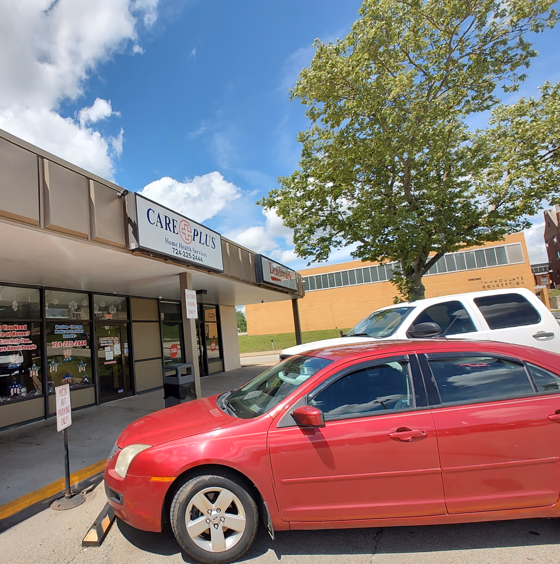 Exterior view of Care Plus Home Health storefront with parked cars and a tree.