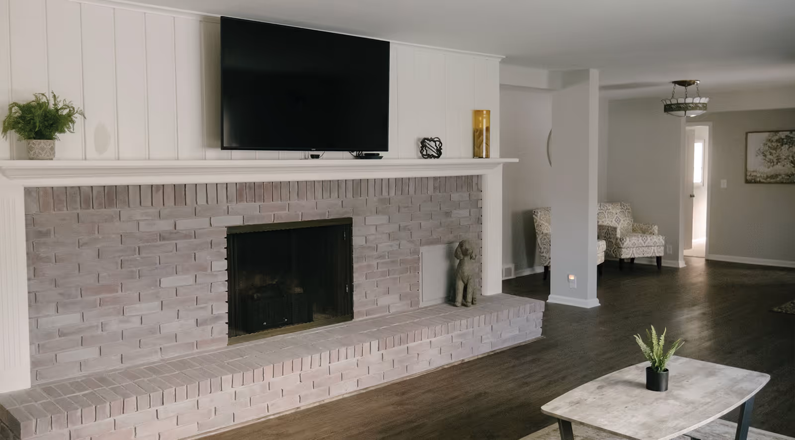Living room with brick fireplace, mounted TV, mantel decor, and seating in background.