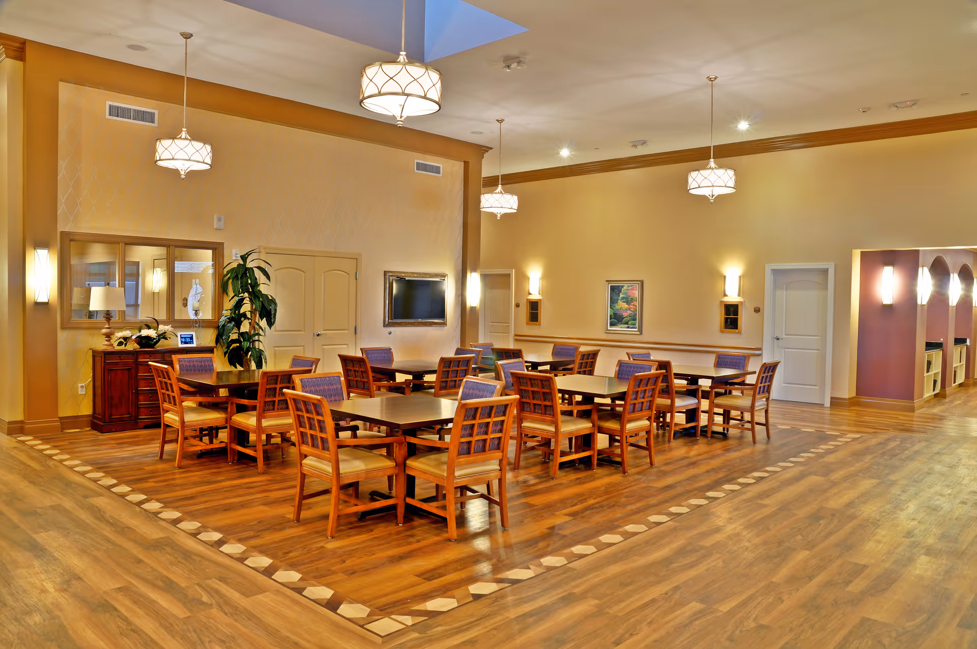 A spacious dining room with multiple wooden tables and chairs arranged neatly on a wooden floor with a decorative border. The room features warm yellow walls, modern pendant lights hanging from the ceiling, a wall-mounted TV, a large mirror, and a plant on a side table. There are several doors and a hallway visible in the background.