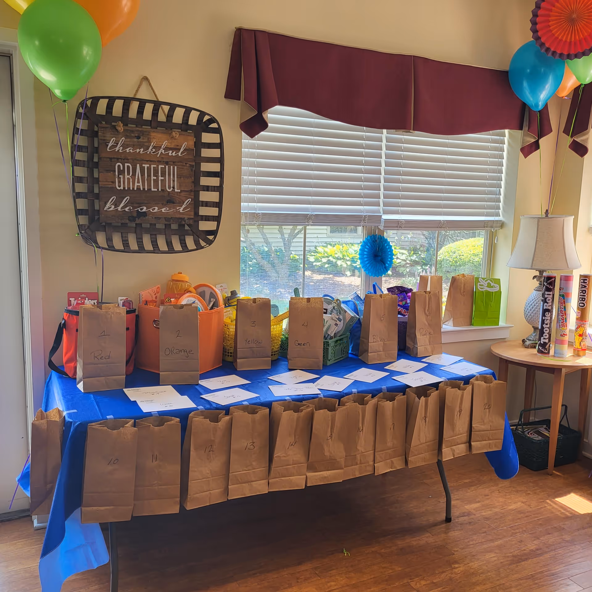 A decorated table with brown paper bags labeled with numbers and colors, set up in front of a window with white blinds. Above the table is a wall decoration with the words 'thankful, grateful, blessed'. Colorful balloons are tied on both sides of the table. A small round table to the right holds large candy containers and a lamp.