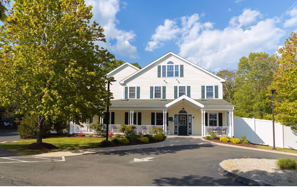 Front exterior view of a two-story white building with green shutters and a covered porch, surrounded by trees and landscaping under a partly cloudy blue sky.