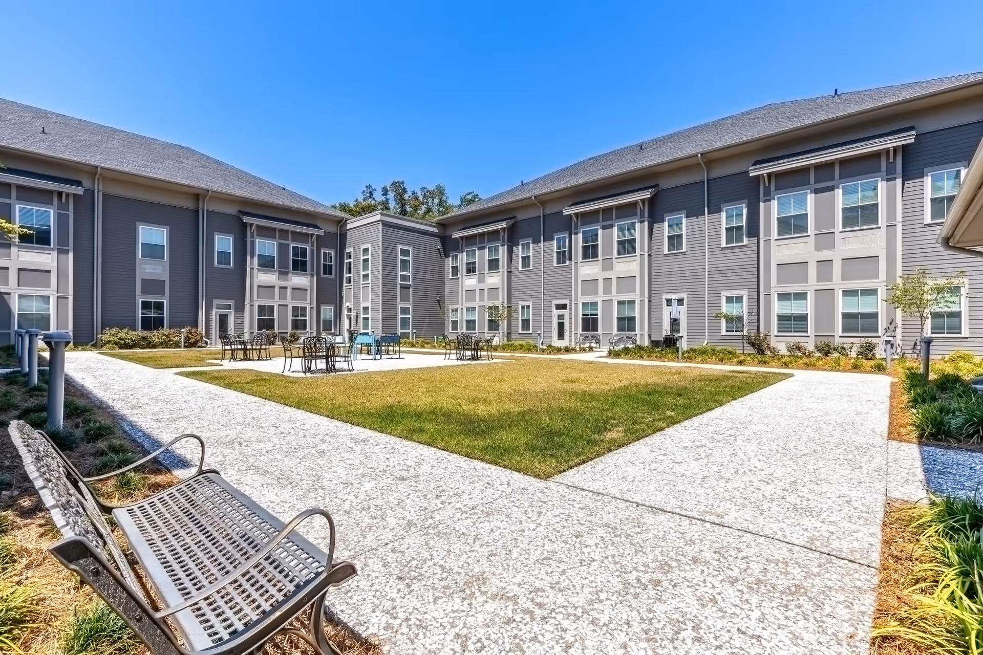 Outdoor courtyard area of Indigo Hall Assisted Living and Memory Care with a grassy center surrounded by concrete walkways, metal benches, and tables with chairs. The courtyard is enclosed by a two-story gray building with multiple windows under a clear blue sky.