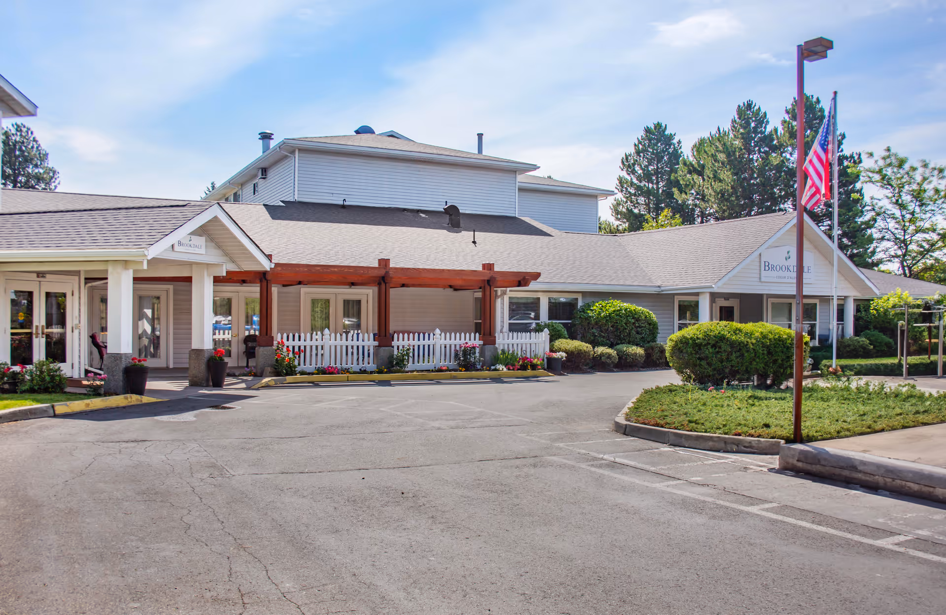 Exterior view of Brookdale Coeur d'Alene senior living facility showing a single-story building with a covered entrance, white picket fence, landscaped bushes, flowers, and an American flag on a flagpole under a clear sky.