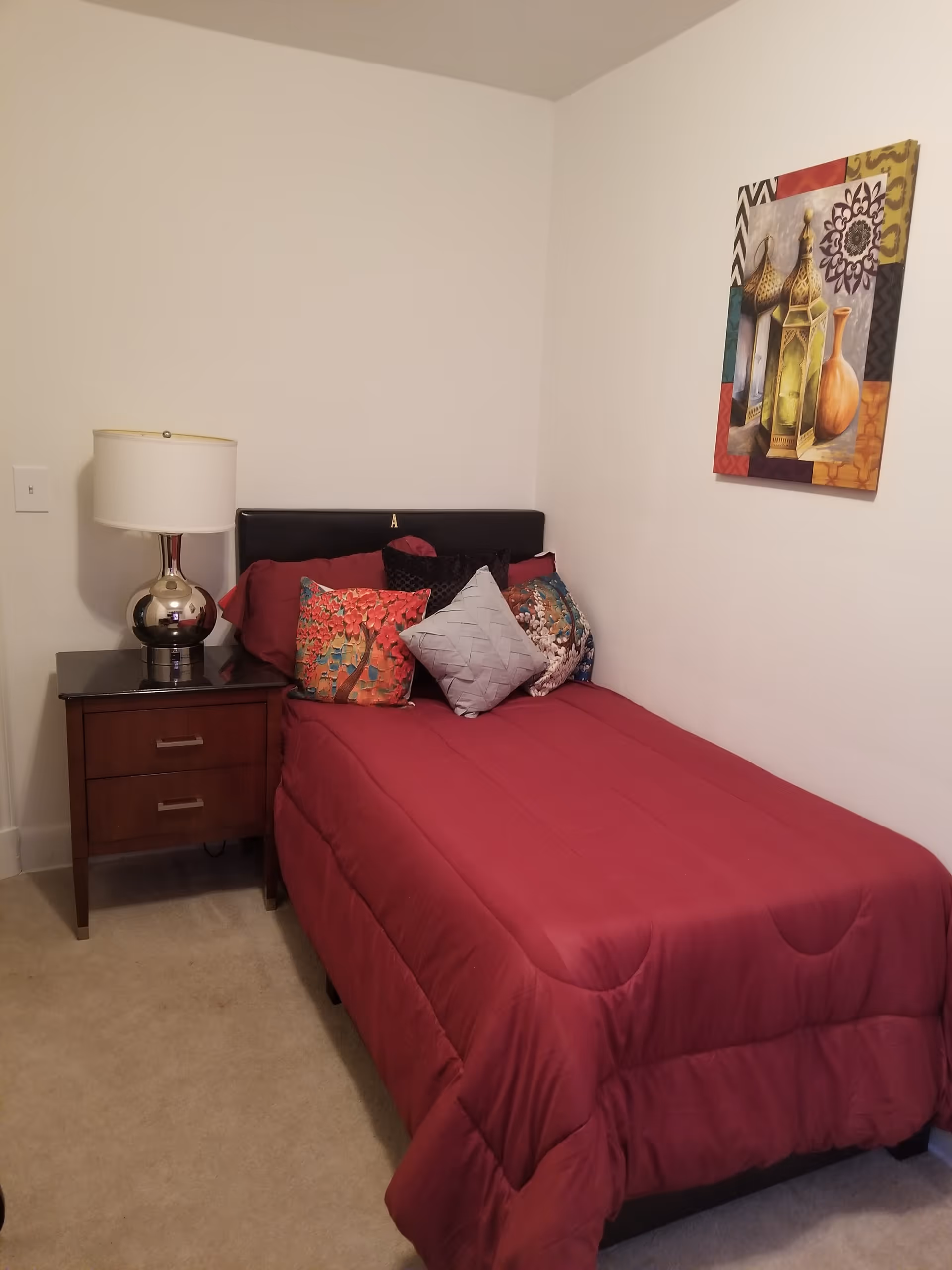 A small bedroom with a single bed covered in a red comforter and multiple decorative pillows. Next to the bed is a wooden nightstand with two drawers and a silver lamp with a white lampshade. A colorful framed artwork hangs on the white wall above the bed.
