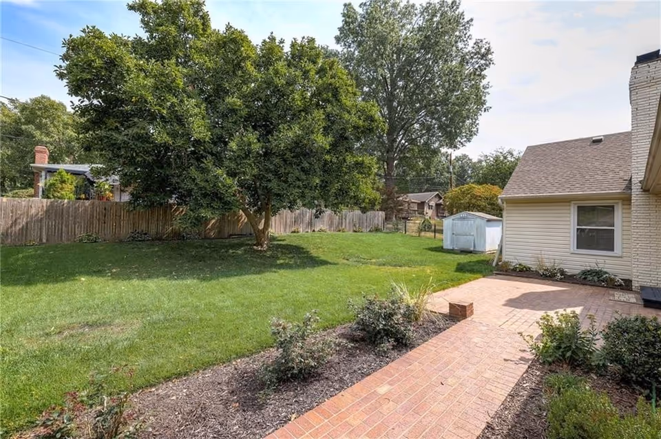 Outdoor backyard of AnnaGrace Senior Living, showing a large tree, green lawn, a brick patio, a wooden fence, and the side of a beige house with a window.