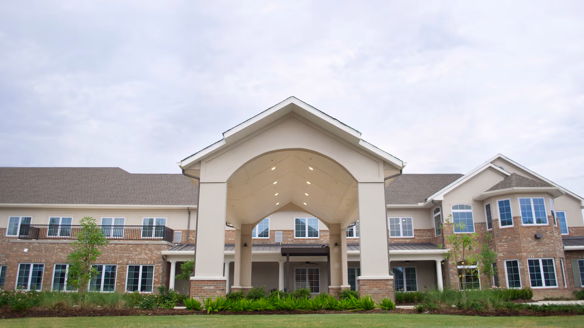 Front exterior view of a two-story senior living facility building with a large covered entrance, multiple windows, and landscaped greenery in front under a cloudy sky.