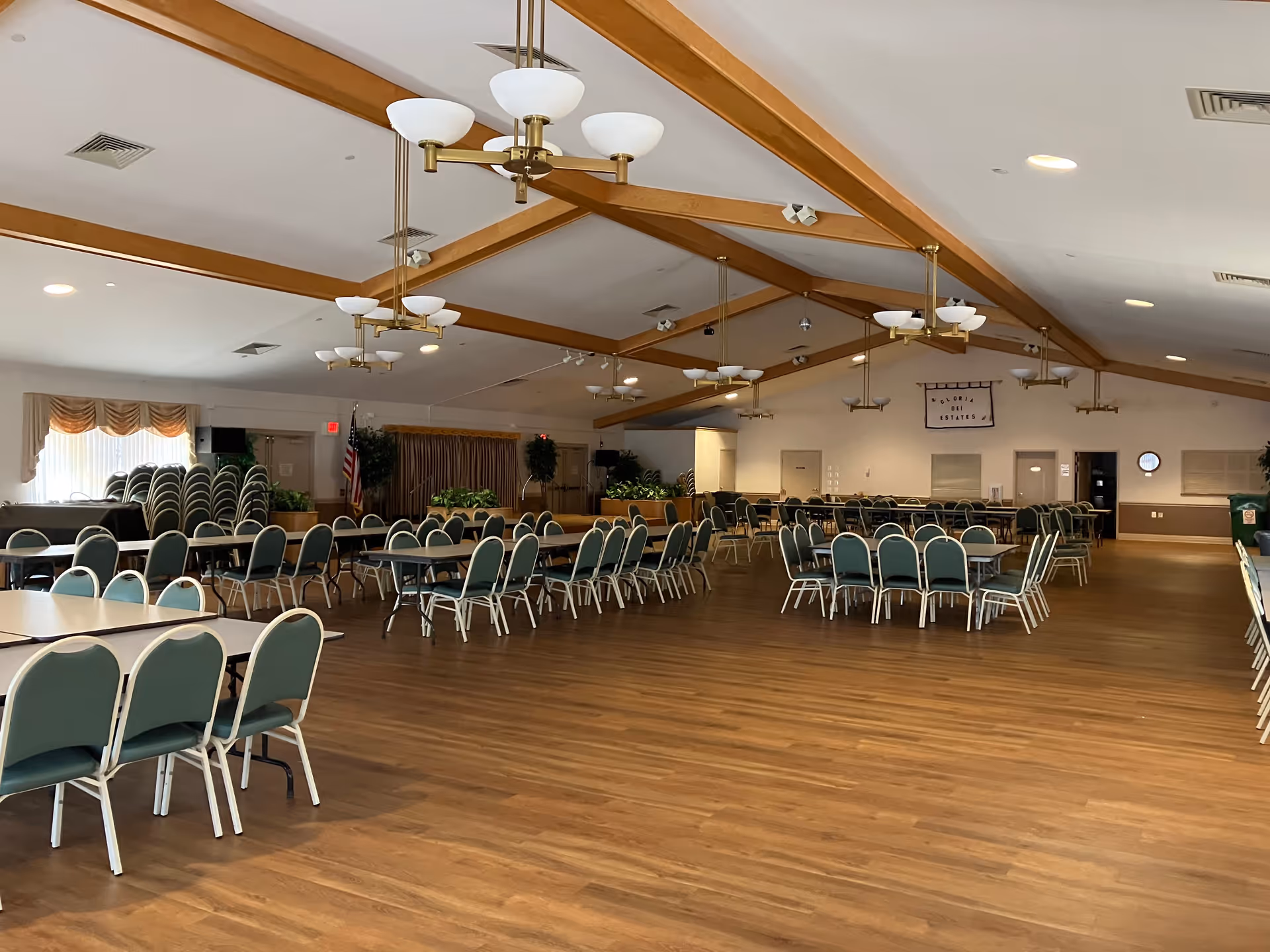 Large multipurpose room with wooden floor and ceiling beams, multiple round and rectangular tables with green cushioned chairs arranged throughout. Several chandeliers hang from the ceiling. Stacked chairs and potted plants are visible near the windows with draped curtains. An American flag stands near the back wall, which has a sign that reads 'Gloria Dei Estates'.