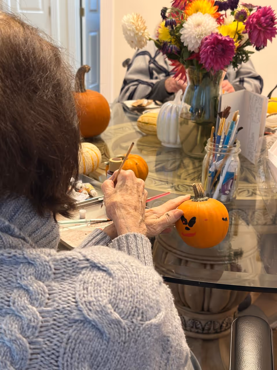 An elderly person wearing a gray knitted sweater is painting a small pumpkin with a face on it at a glass table. There are other small pumpkins, paintbrushes in a jar, and a vase with colorful flowers on the table. Another person is partially visible in the background.