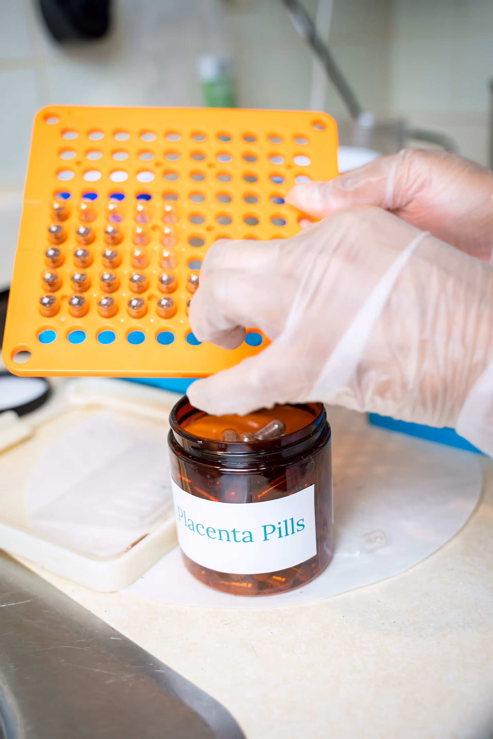 Gloved hands handle an orange perforated tray of capsules beside a labeled placenta pills jar.