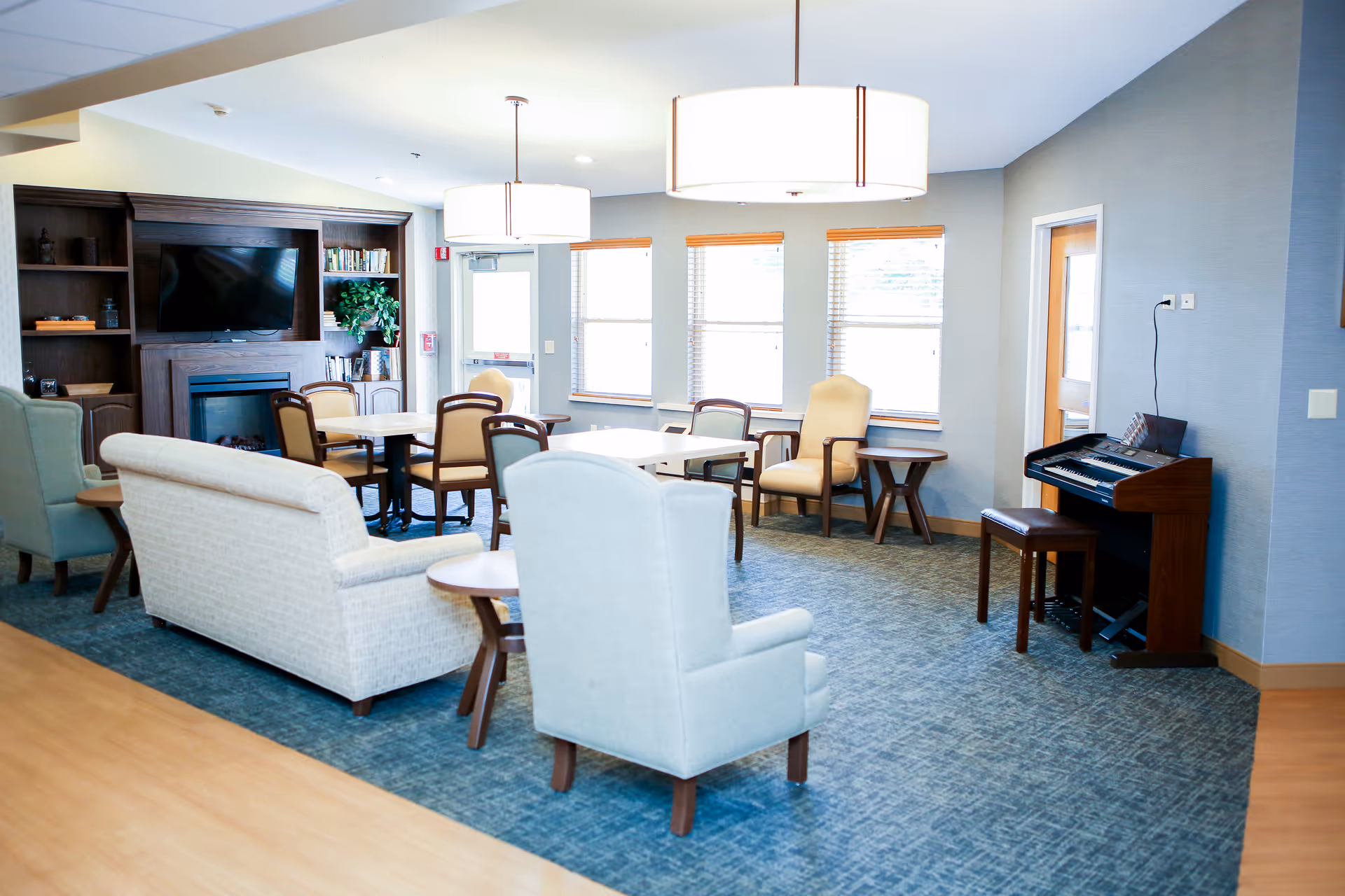 Bright senior living lounge with sofas, armchairs, tables, piano, and TV near fireplace.