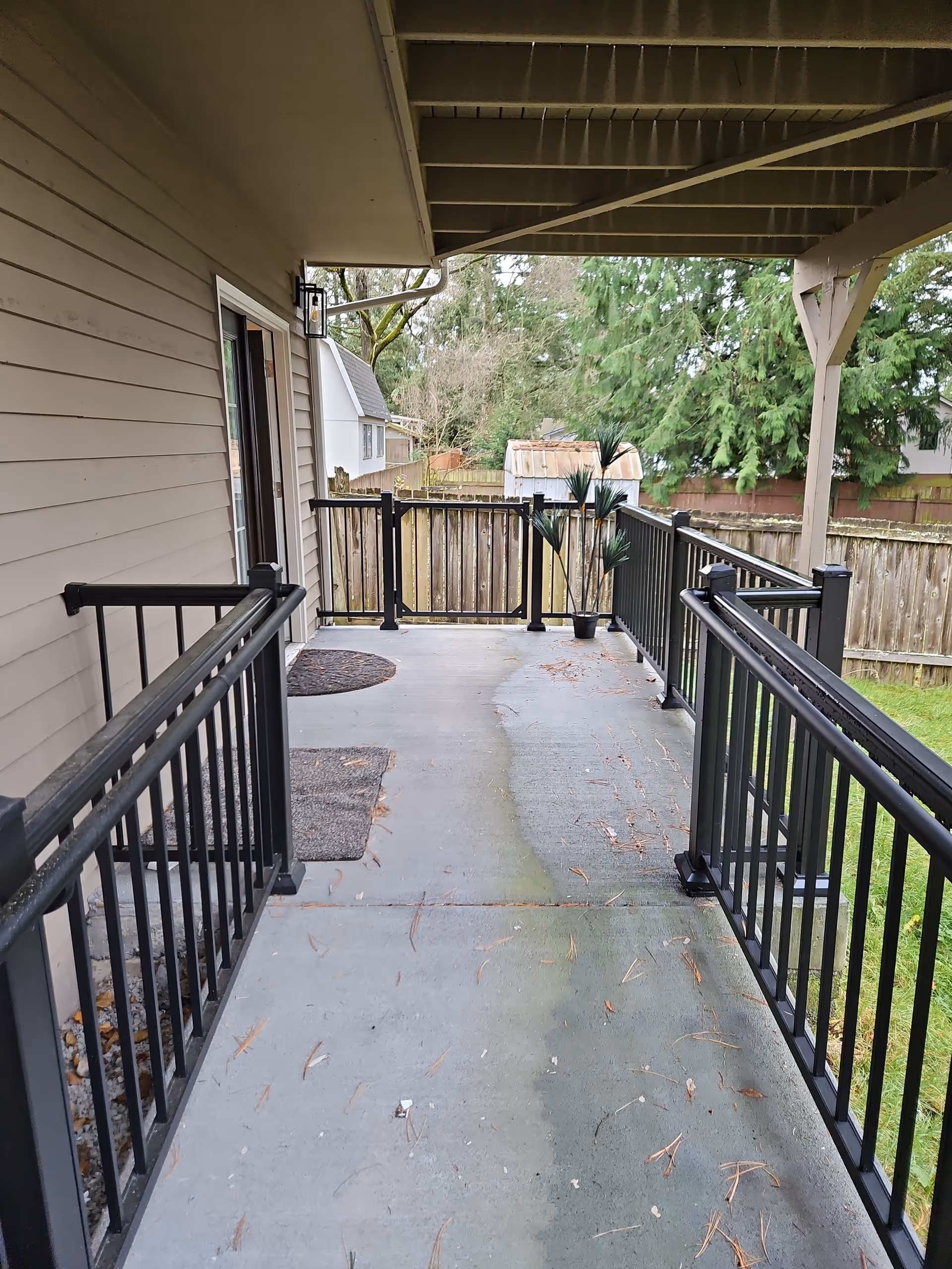 Covered outdoor deck with black railings, sliding door, and trees beyond.