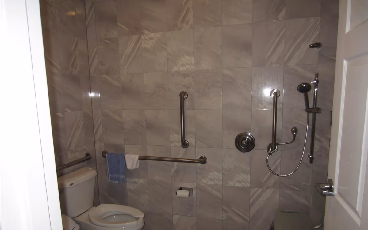 Bathroom with gray tiled walls, a toilet with grab bars on the side and back walls, a mounted showerhead with adjustable height, and a towel hanging on one of the grab bars.