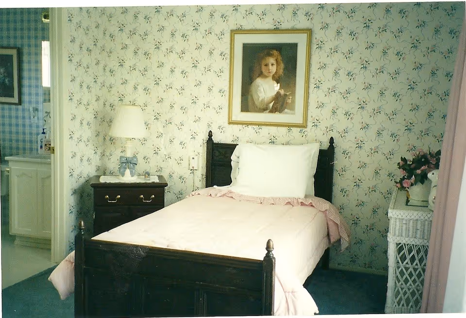 A cozy bedroom with floral wallpaper, dark wood bed, lamp, and framed portrait.