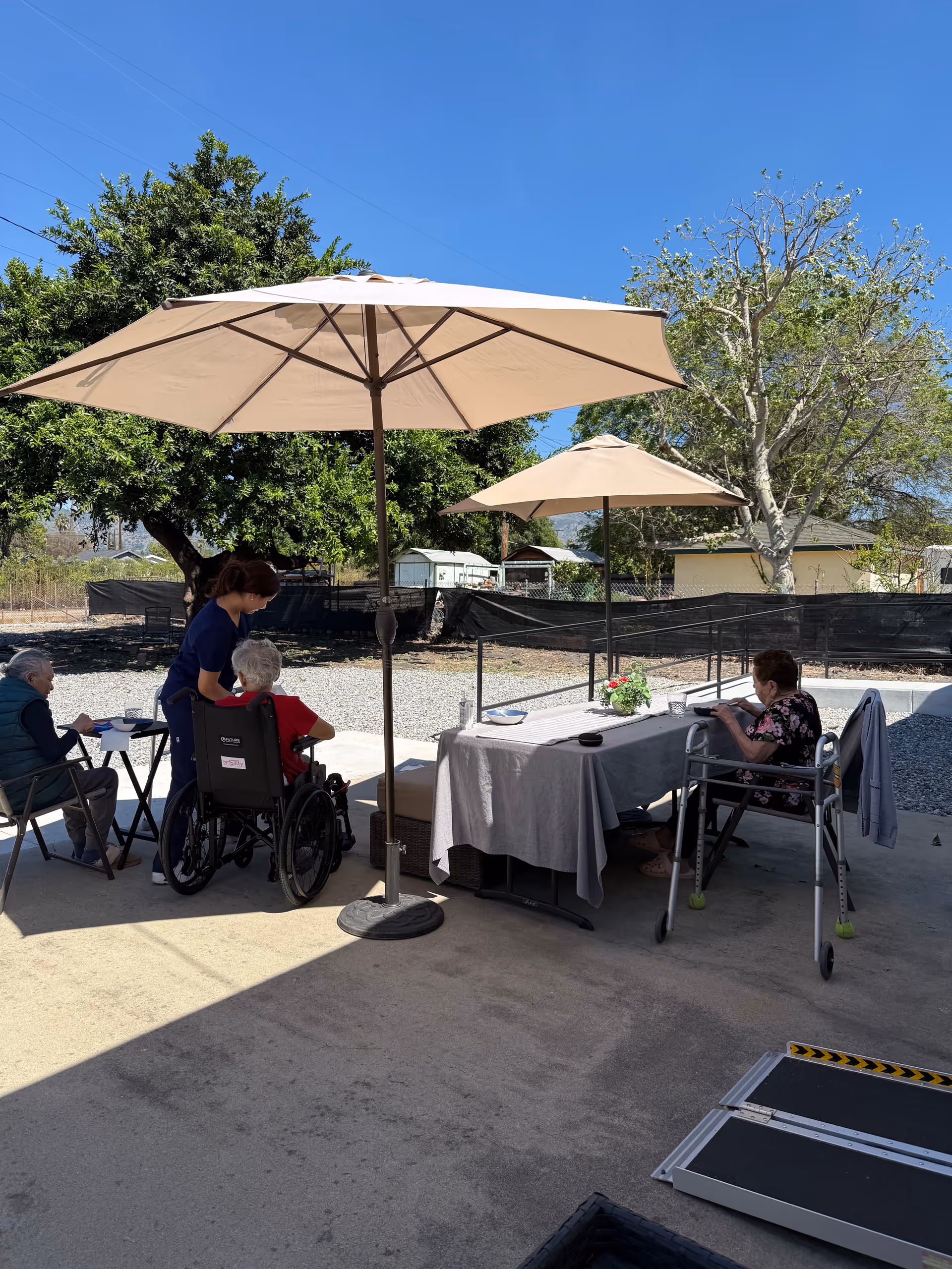 Outdoor patio with seniors in wheelchairs under umbrellas, caregiver assisting at a table.