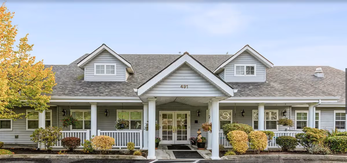 Single-story building front with a covered entrance porch supported by columns, dormer windows, and landscaped shrubs.