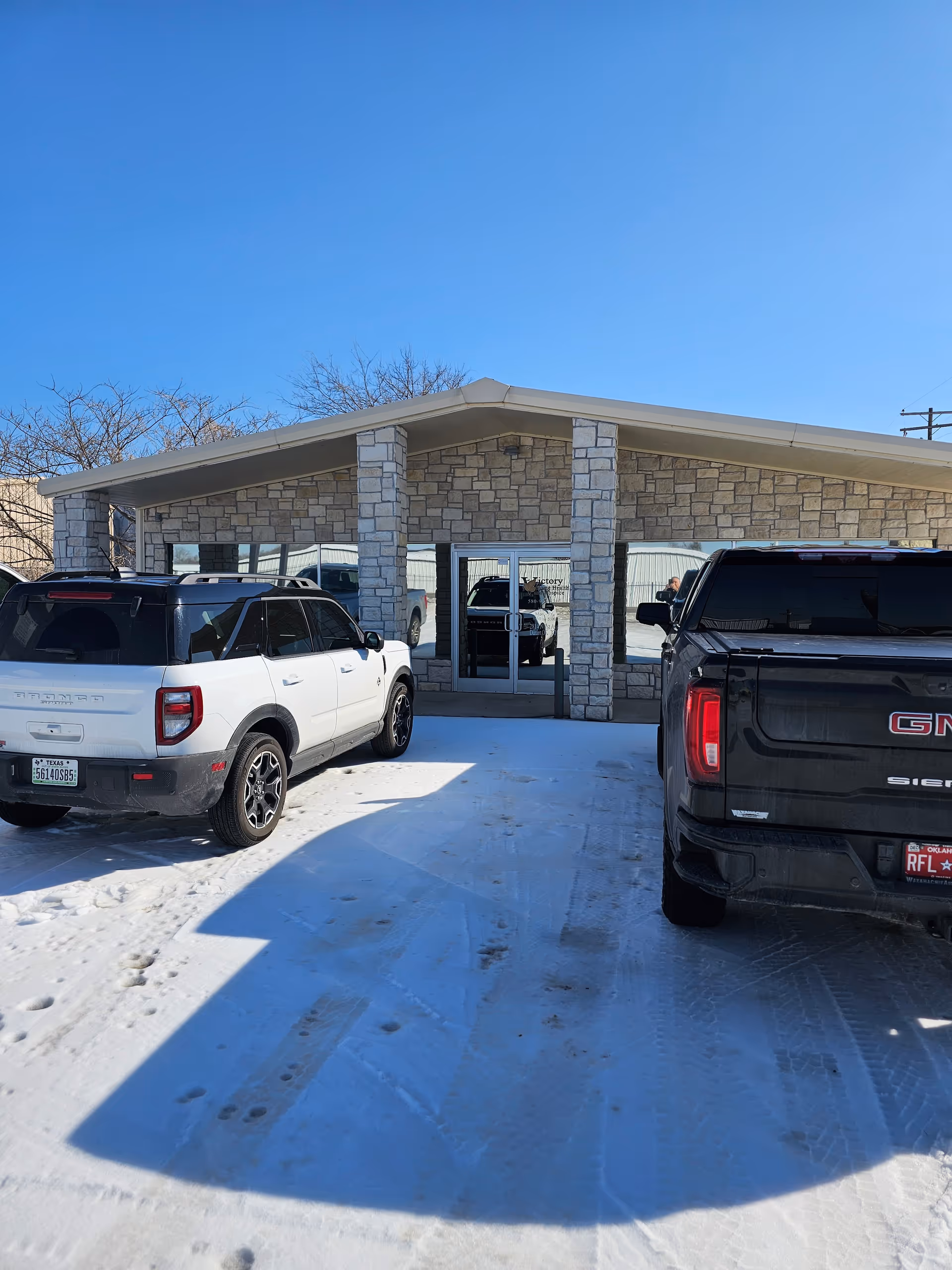 Stone-faced building entrance with glass doors, snow-covered parking lot, two parked SUVs.