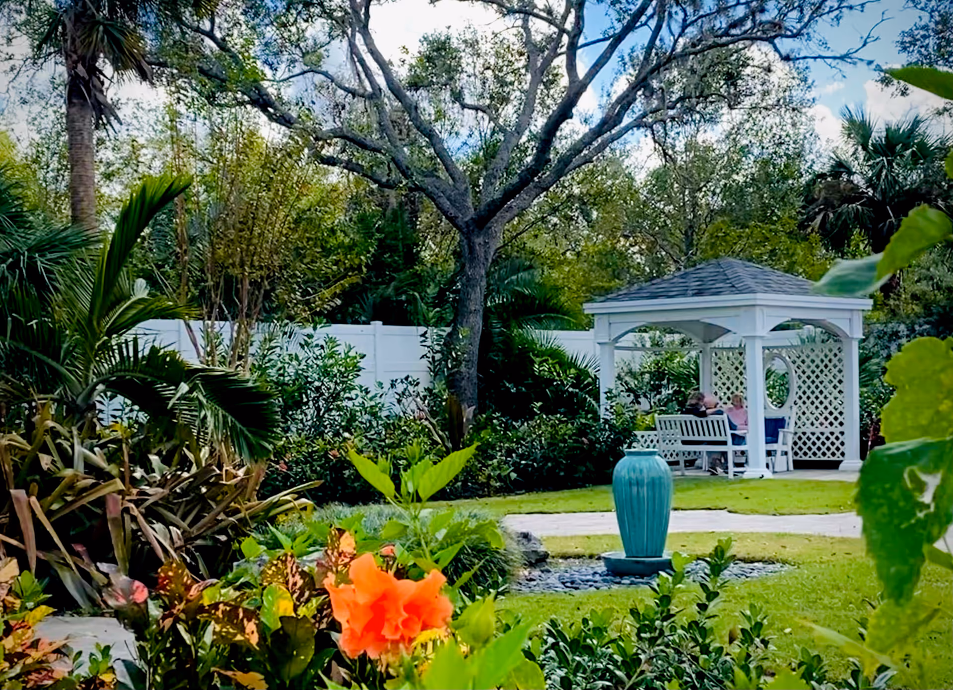 A serene garden with a white gazebo, benches, a turquoise fountain, and lush tropical plants.