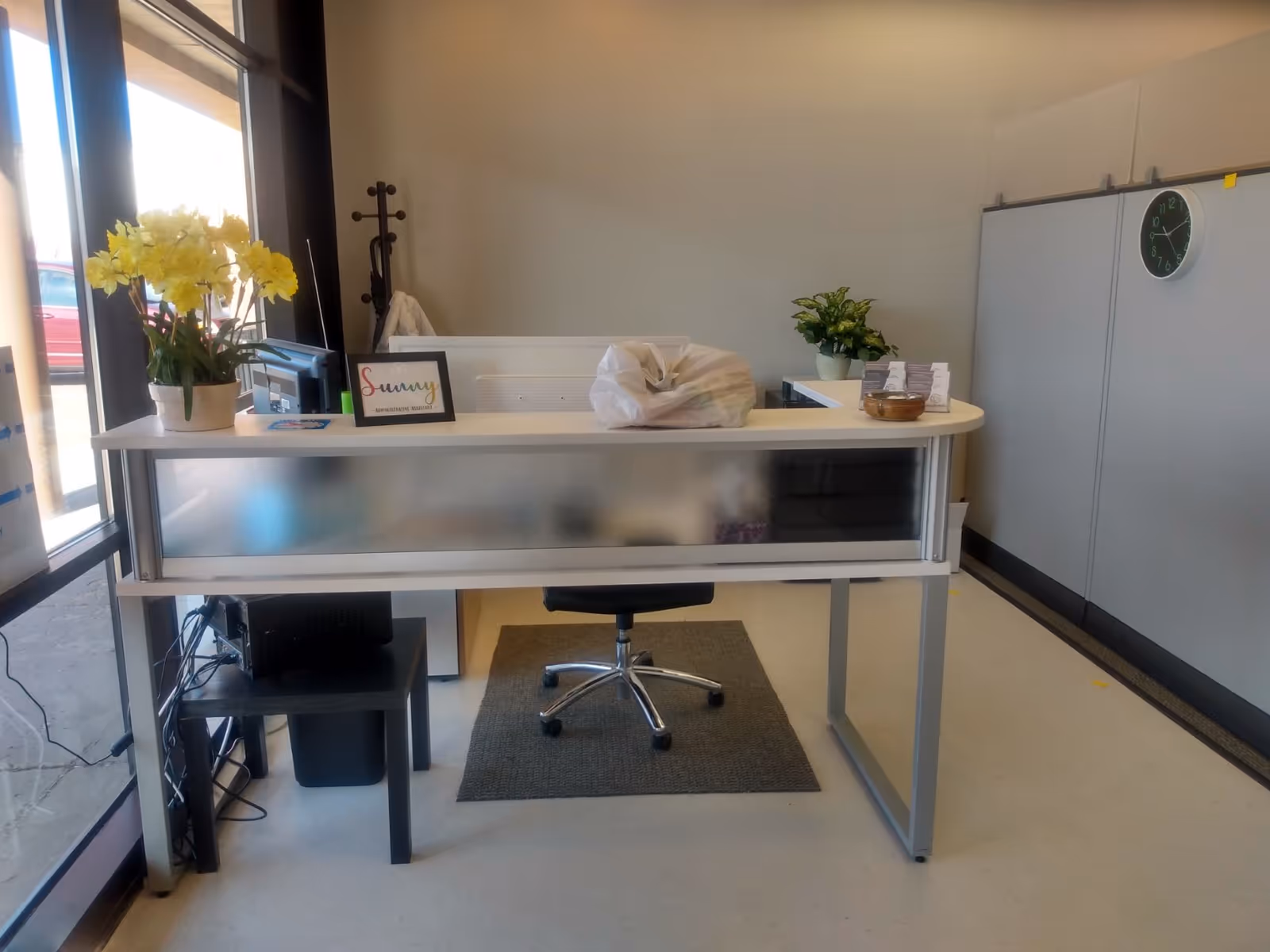 Reception desk in a bright facility lobby with flowers, plants, and office equipment.