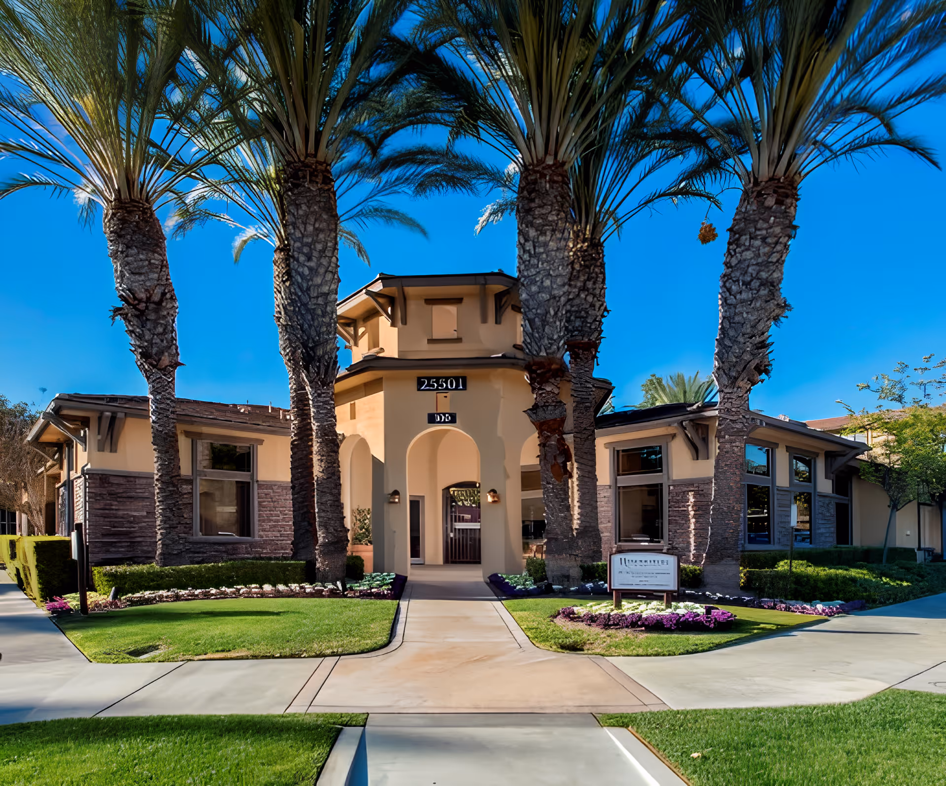 Front entrance of a beige single-story building with arched entry, flanked by tall palm trees, manicured lawns, and a paved walkway.