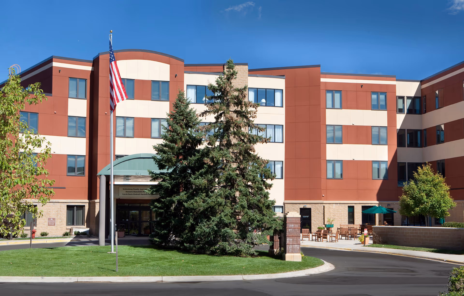 Four-story red and beige senior living facility with entrance canopy, flagpole, and outdoor seating.