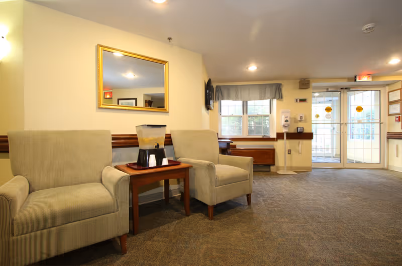 A waiting area with two beige armchairs separated by a wooden side table holding a beverage dispenser and cups. A large rectangular mirror hangs on the wall above the chairs. The room has carpeted flooring, a window with a blue valance, and double glass doors leading outside. The walls are painted light yellow, and there are ceiling lights illuminating the space.