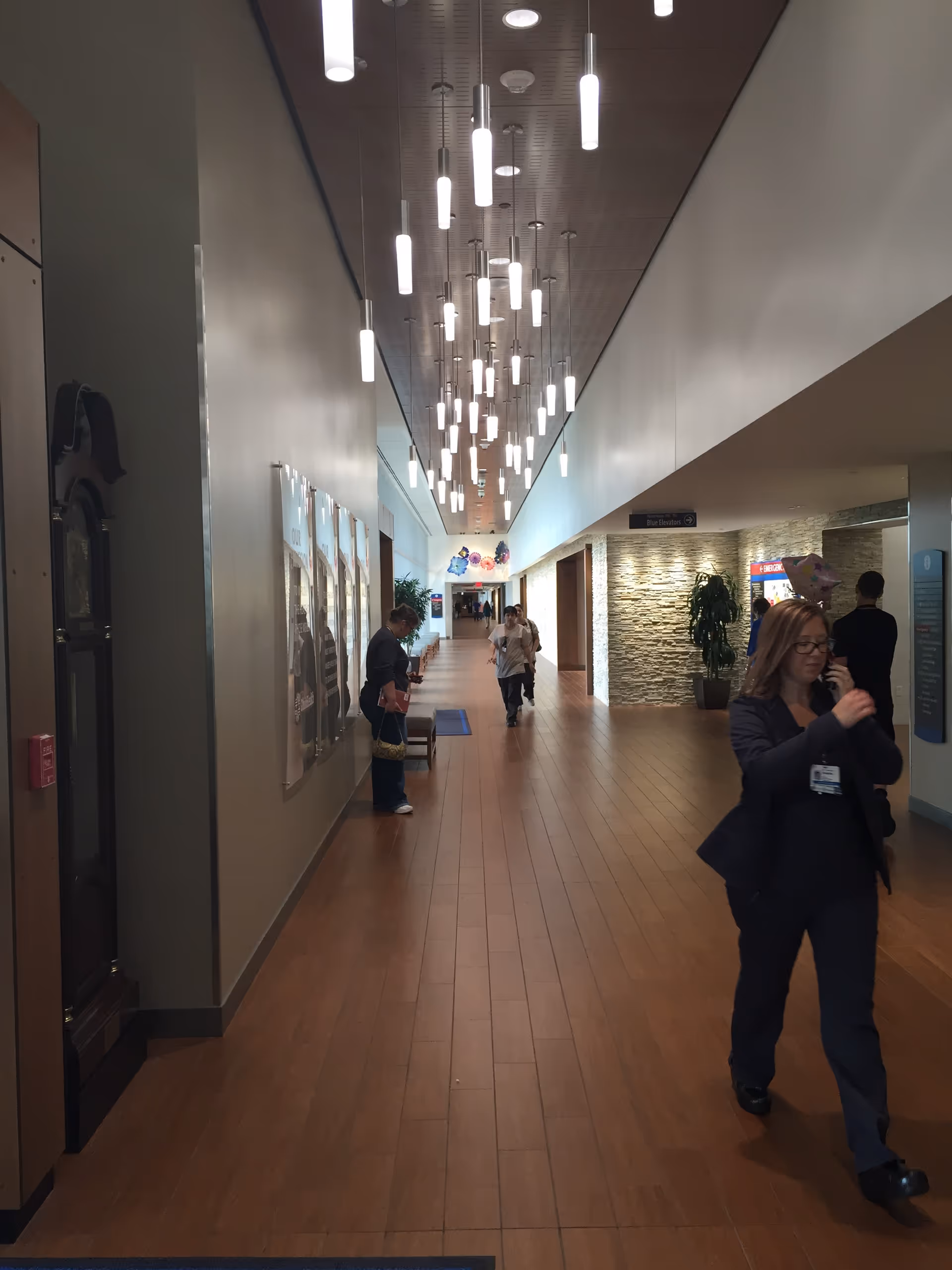 Long, well lit corridor in a medical facility with hanging lights and staff walking.