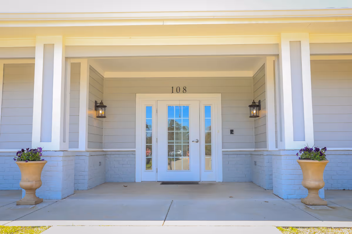 Front entrance of a light-gray building with double glass doors numbered 108, flanked by wall lanterns and flower urns.