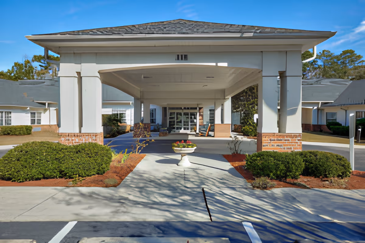Front entrance of TerraBella Summerville facility featuring a covered driveway with white columns and a flower planter in the center, surrounded by bushes and a clear blue sky.