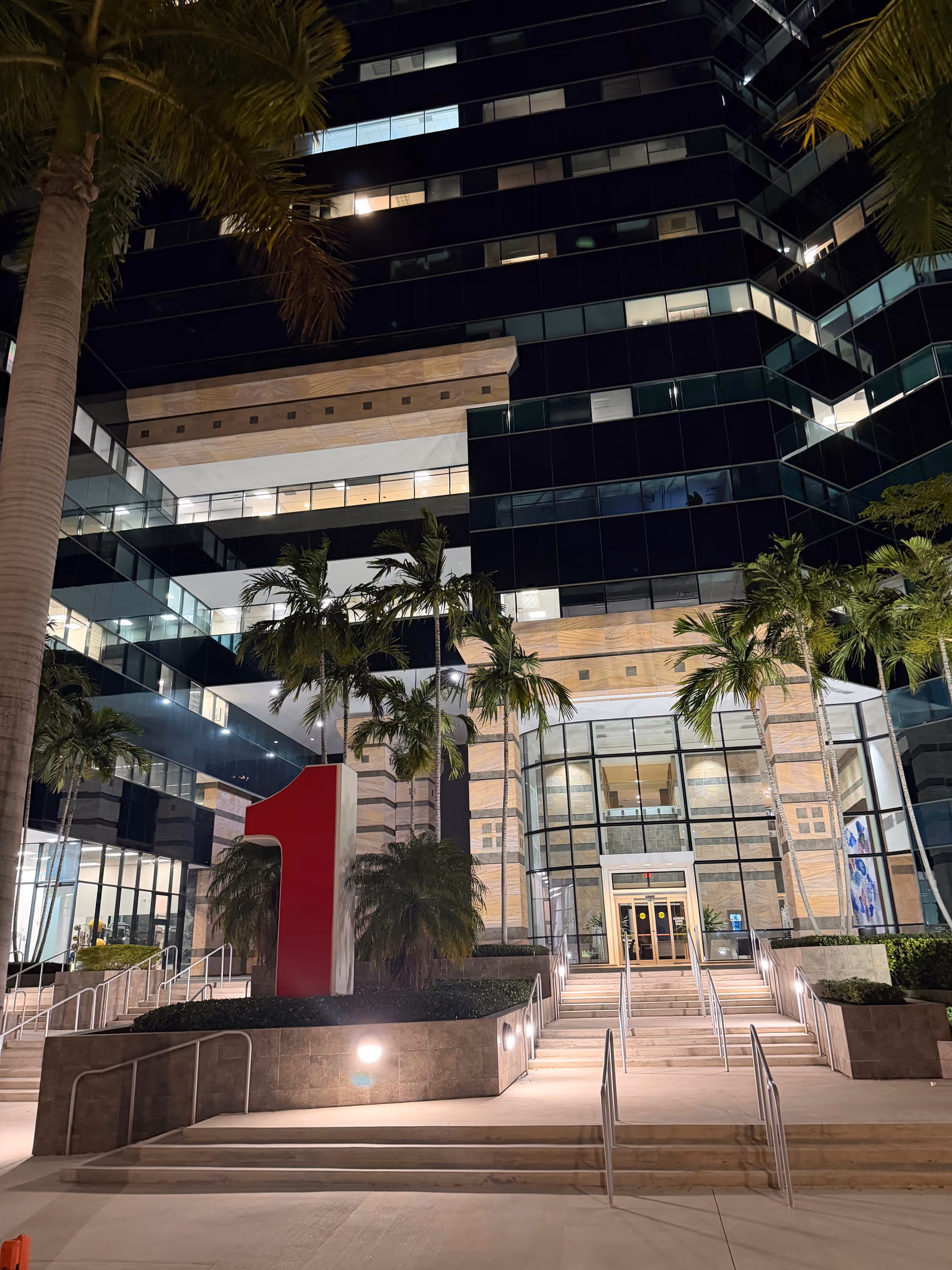 Night view of a modern building entrance with palm trees and a large red numeral sculpture.