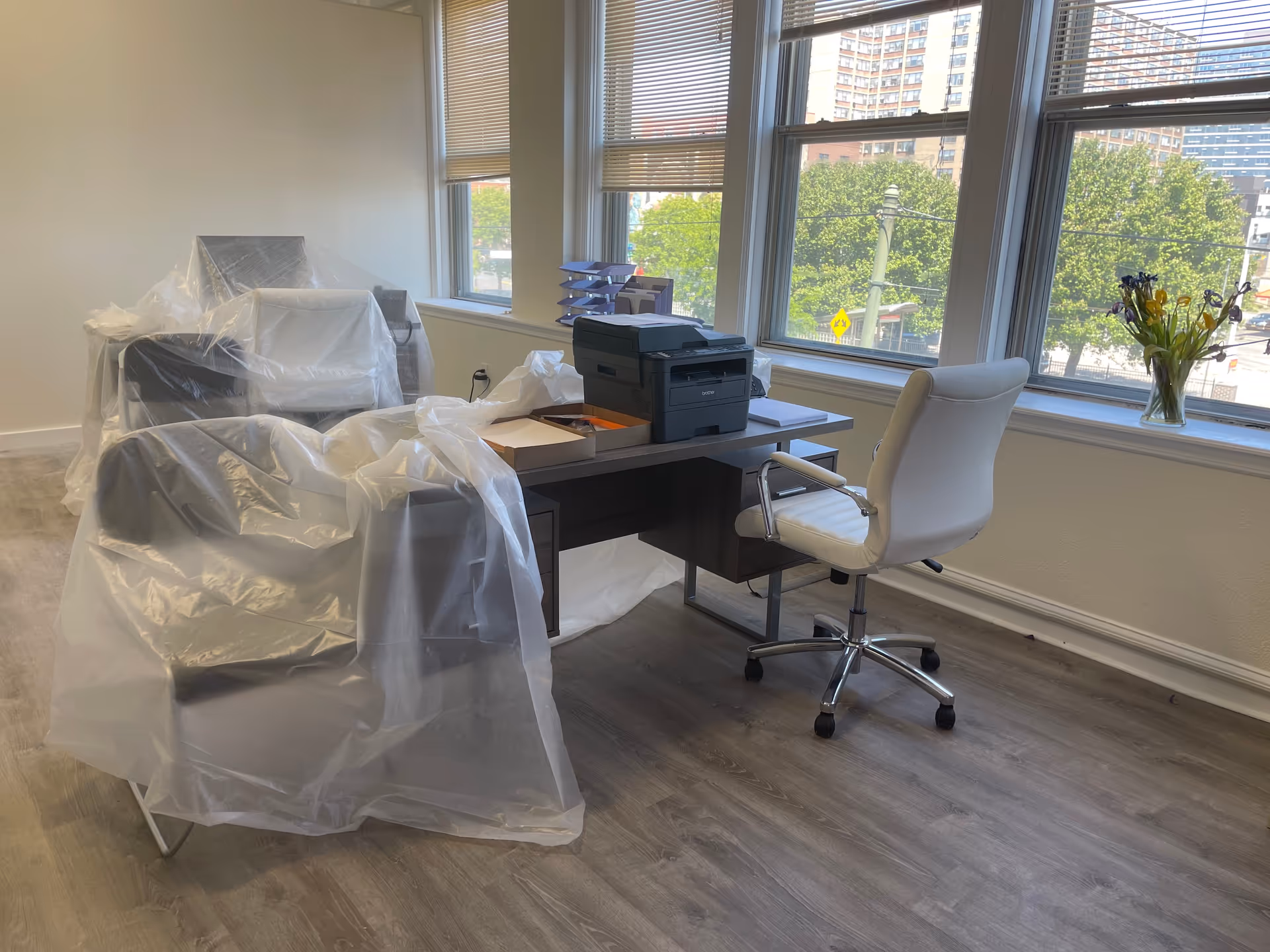 Sunlit office area with plastic-wrapped furniture and a desk by large windows.