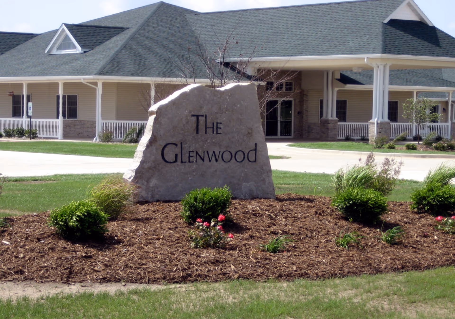 Large stone sign with the words 'The Glenwood' engraved on it, placed in a landscaped area with bushes and flowers in front of a single-story building with a green roof and white columns.