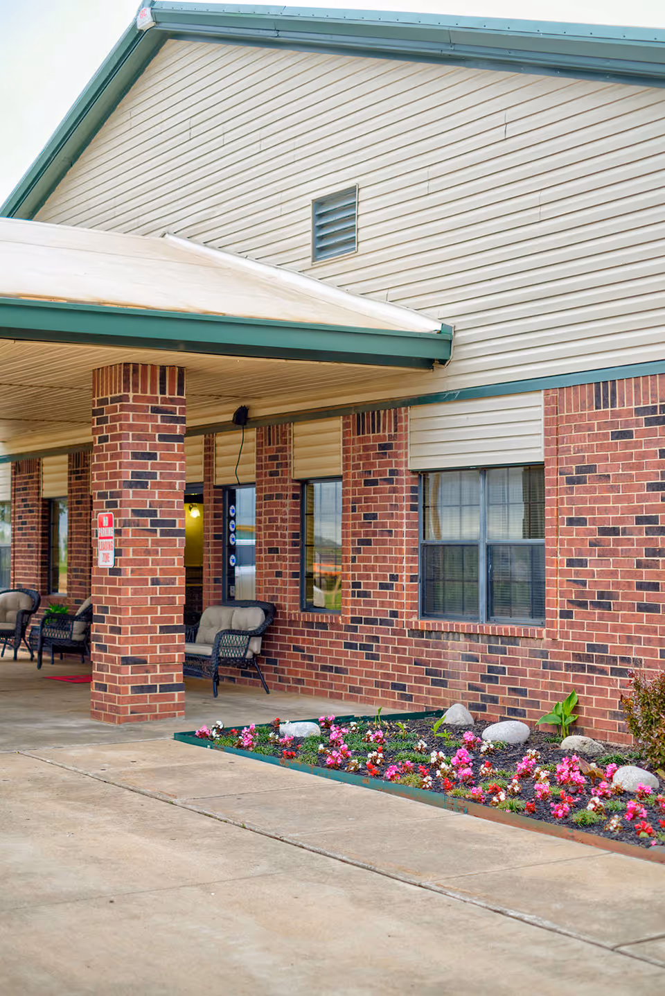Exterior view of a brick building with a covered entrance supported by brick columns. There are cushioned chairs placed under the covered area and a flower bed with pink and red flowers along the sidewalk.