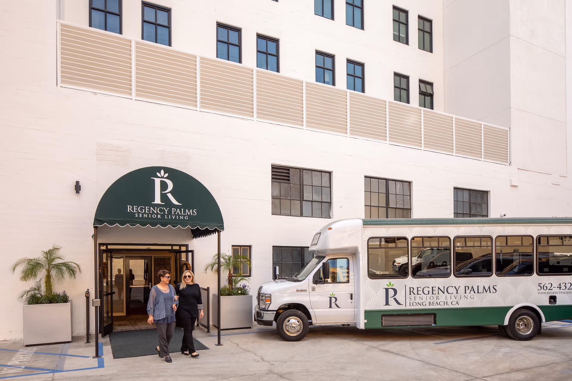 Two women walking out of the entrance of Regency Palms Senior Living facility. The entrance has a green awning with the Regency Palms logo and name. A white and green shuttle bus with the Regency Palms logo and contact information is parked nearby. The building is white with multiple windows.