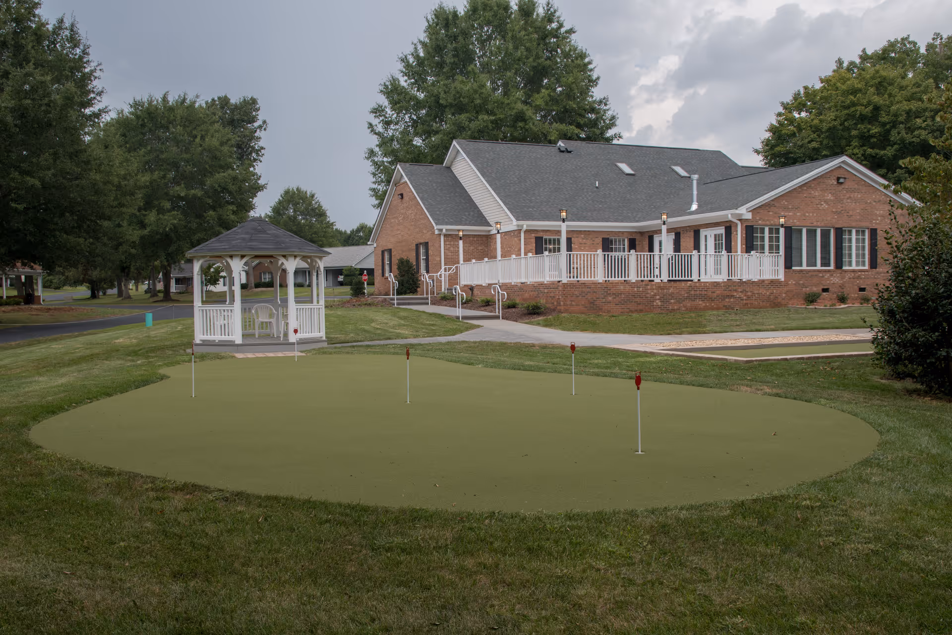 Brick retirement community building with a white gazebo and a small putting green in the foreground under a cloudy sky.