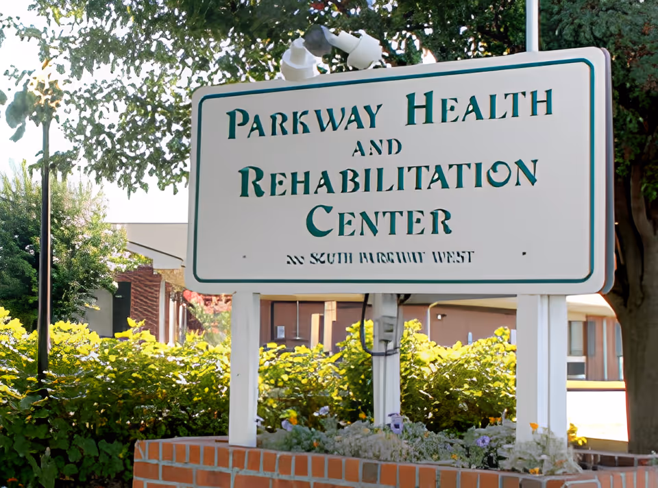Outdoor sign for Parkway Health and Rehabilitation Center surrounded by greenery and flowers, with part of the building visible in the background.