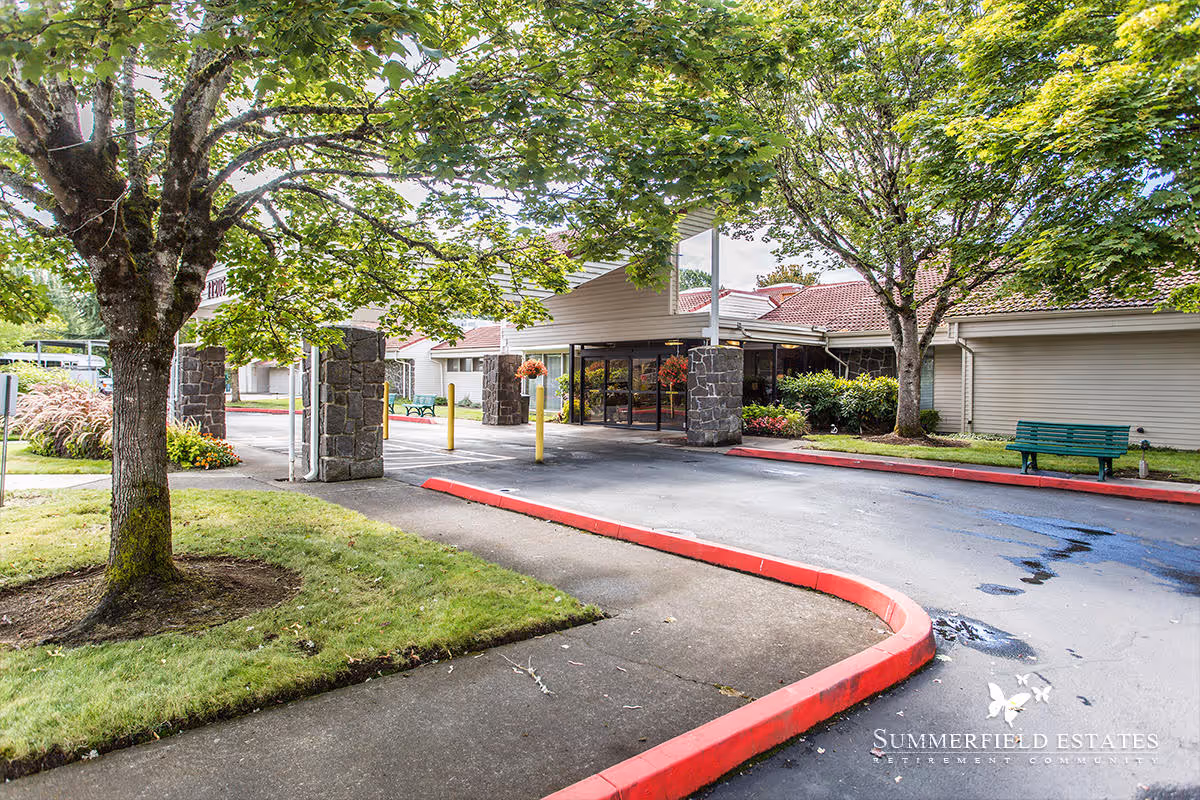 Entrance of Summerfield Estates Retirement Community showing a covered driveway with stone pillars, surrounded by green trees and landscaped grass areas, with a green bench on the right side.