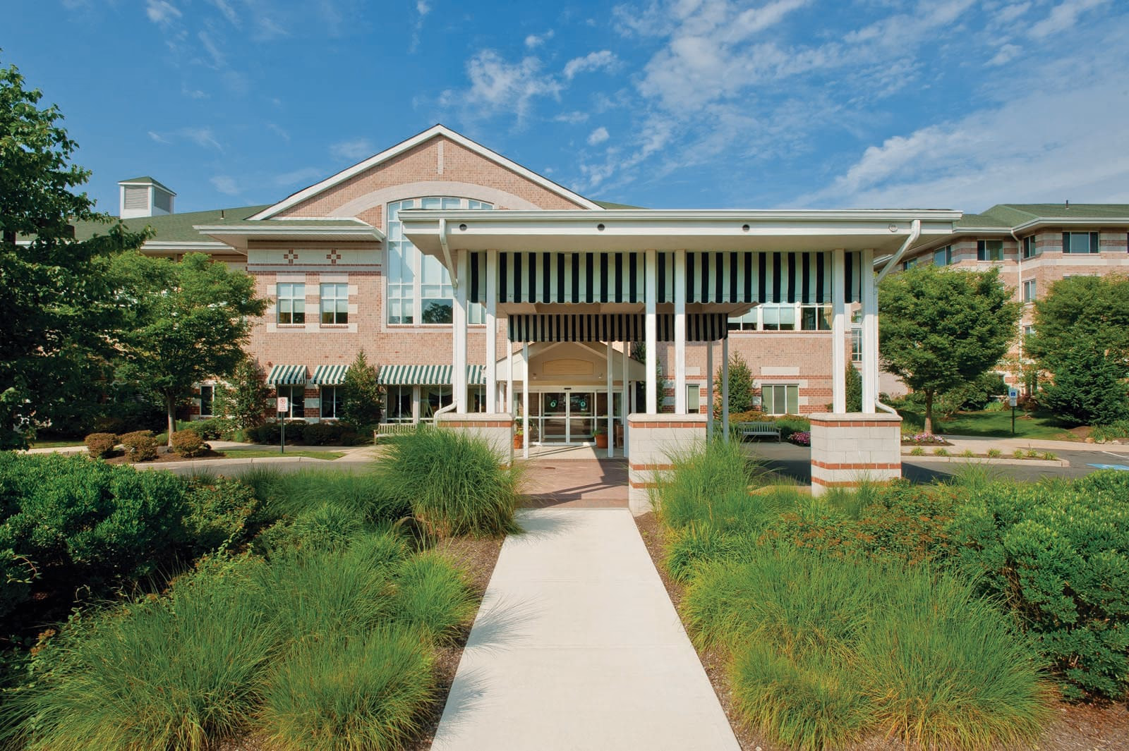Front exterior view of a large brick building with a covered entrance featuring black and white striped awnings. The building is surrounded by well-maintained greenery and trees under a blue sky with some clouds.