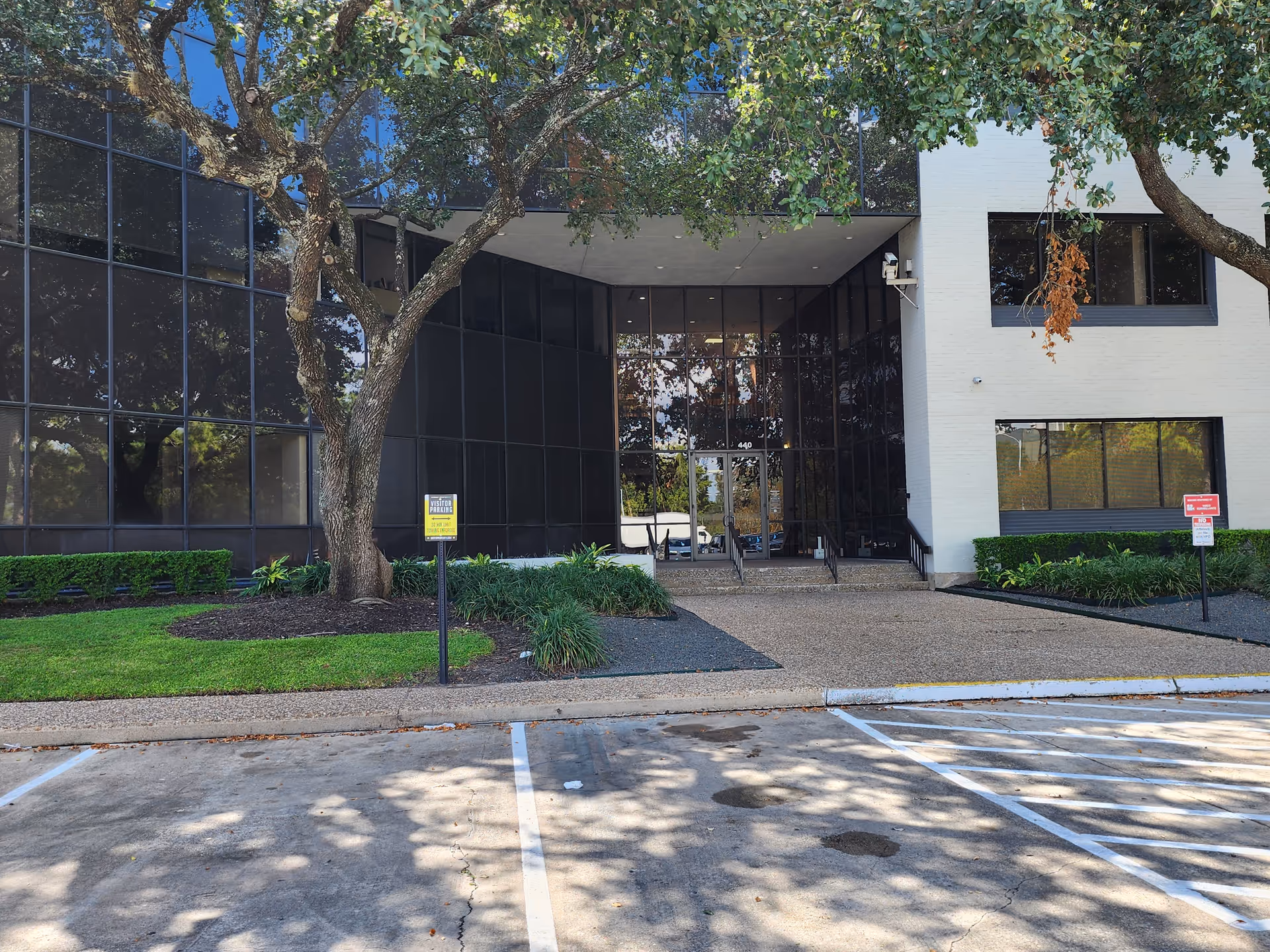 Modern building entrance with glass walls, trees, and a paved walkway.
