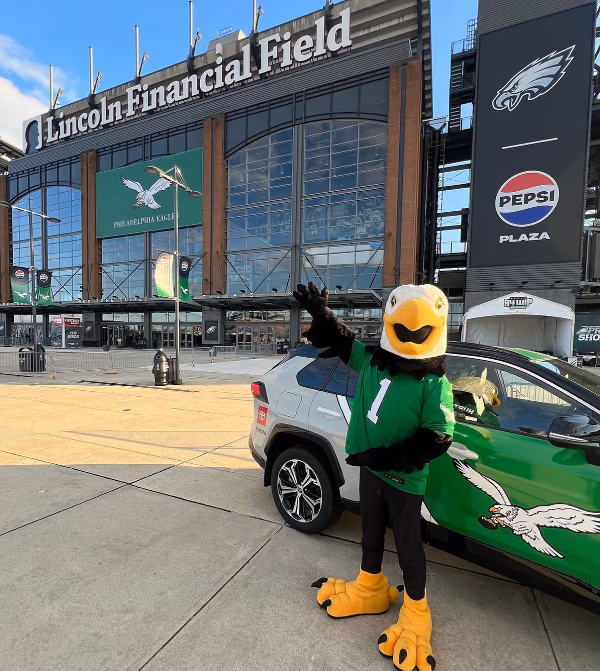 Costumed eagle mascot in green jersey waves beside a car outside Lincoln Financial Field.