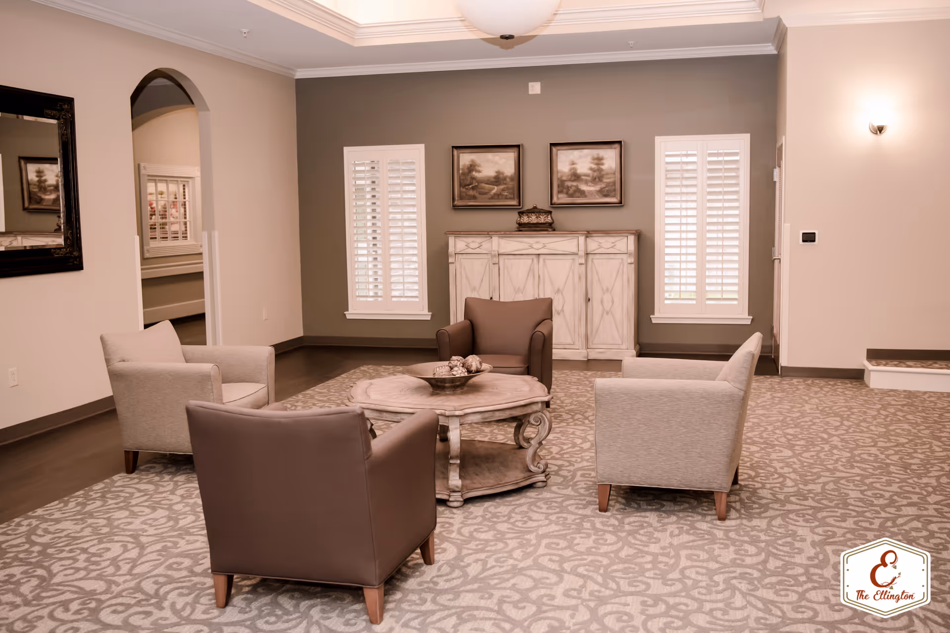 A cozy sitting area with four armchairs arranged around a round wooden coffee table on a patterned carpet. The room has two windows with white shutters, a white wooden cabinet against a gray accent wall, two framed paintings above the cabinet, and a large mirror on the left wall. Soft lighting illuminates the space.