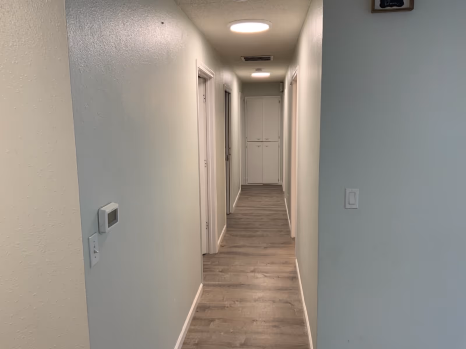 Long, well-lit hallway with light blue walls, doors on both sides, and a cabinet at end.
