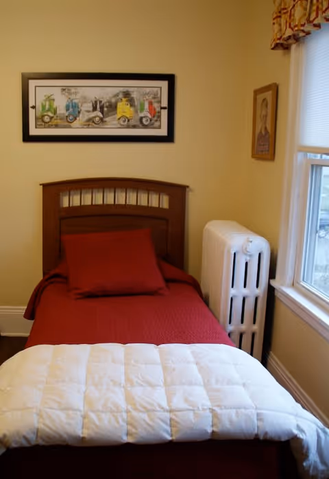 A small bedroom with a single bed covered with a red blanket and a white quilt folded at the foot. There is a red pillow on the bed. The headboard is wooden with a slatted design. On the wall above the bed is a framed picture of colorful vintage cars. To the right of the bed is a white radiator and a window with blinds and a patterned valance. A small framed portrait hangs on the wall next to the window.