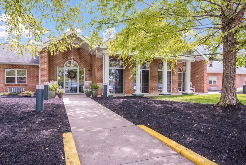 Brick senior living facility entrance with columns, walkway, and landscaping.