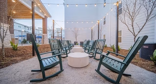 Outdoor courtyard with green wooden rocking chairs arranged around round concrete tables under string lights between buildings.